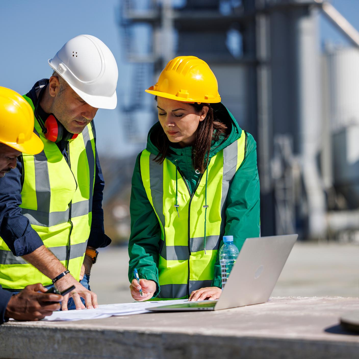 Trois ingénieurs en tenue de sécurité discutent dans une installation industrielle en écrivant sur un papier à côté d’un ordinateur portable.