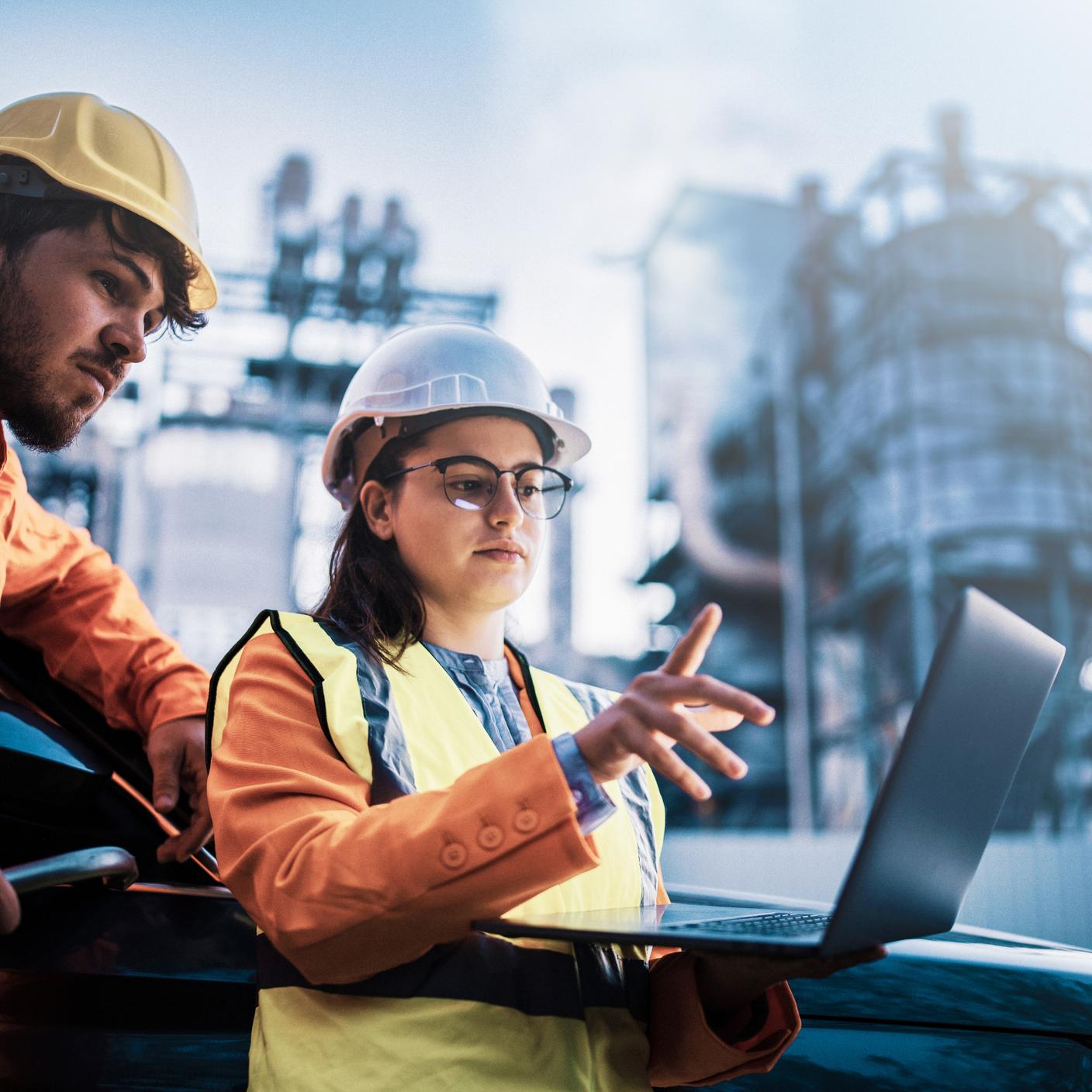 Dos asociados vestidos de seguridad revisan datos en una computadora portátil frente a una planta de energía.